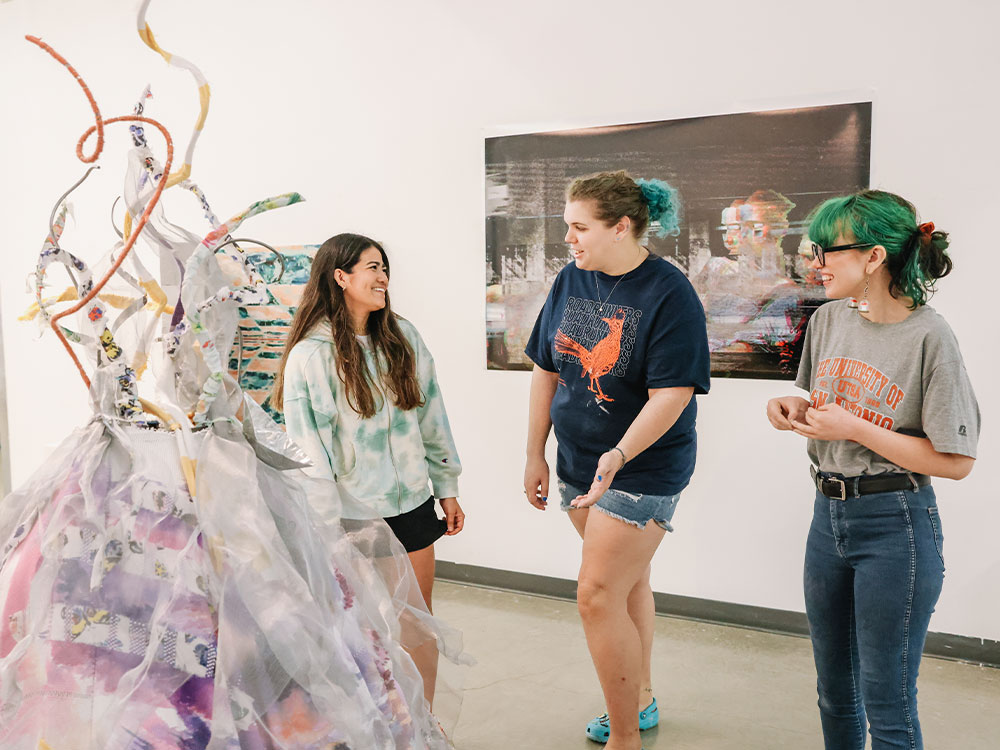 Three UTSA students examining and discussing a mixed media art sculpture.