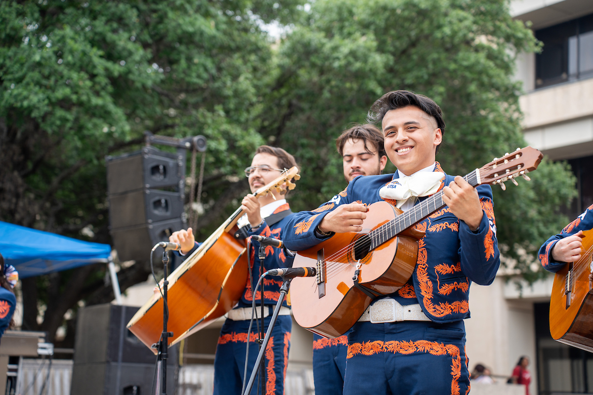 A student mariachi group performs outdoors, with musicians dressed in traditional embroidered charro outfits playing guitars and other string instruments.