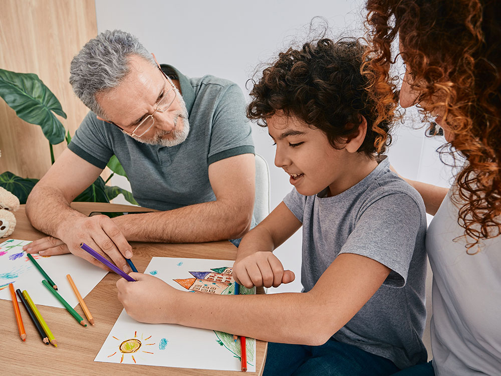 A young boy sitting in his mother's lap drawing a picture with an art therapist looking over his work.