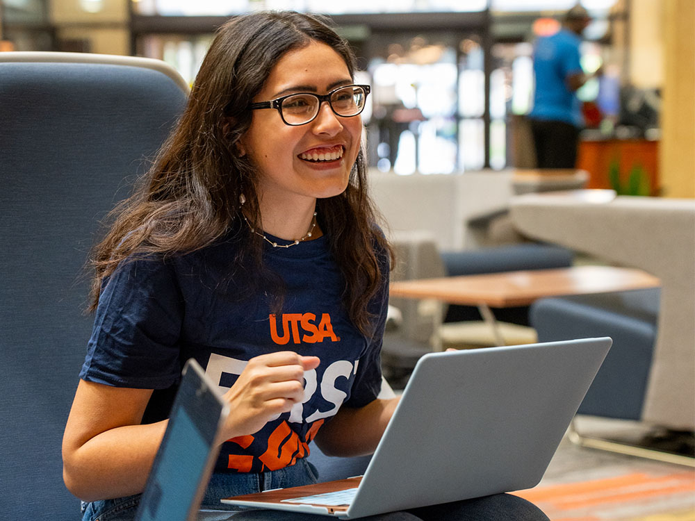 A student wearing a UTSA shirt works on a laptop inside the John Peace Library.