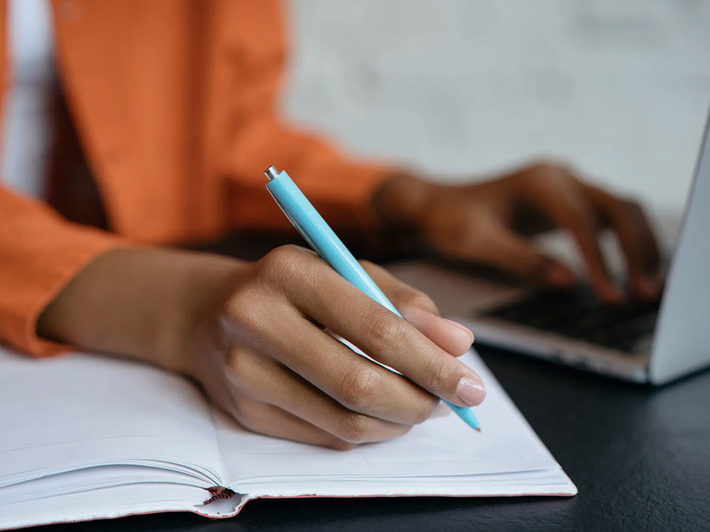 Person taking notes in a notebook while using laptop.